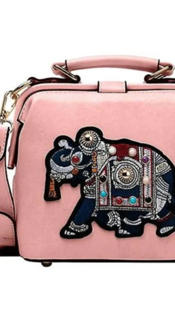 A close-up product photograph from 4_8.webp showing a dusty pink faux-leather crossbody handbag featuring a large, ornate embroidered elephant patch on the front. The elephant is depicted in profile with detailed dark blue and white stitching, wearing a decorative blanket or caparison filled with geometric patterns, white swirls, a circular mandala motif, and colorful stud embellishments in red, turquoise, purple, and gold. The structured bag includes a pink top handle, gold metal hardware hardware rings, and a matching shoulder strap.