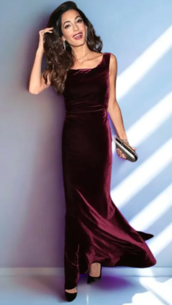 A full-length evening fashion photograph from 4_9.webp of a woman with long, voluminous brown waves posing against a light blue wall with diagonal shadow highlights. She is dressed for a formal occasion, wearing a floor-length, sleeveless gown made of luxurious deep burgundy velvet. She carries a small, structured oval hard-shell box clutch with a metallic bronze sheen in one hand and pairs the dress with black pointed-toe heels, illustrating an effortlessly chic jewel-toned outfit.