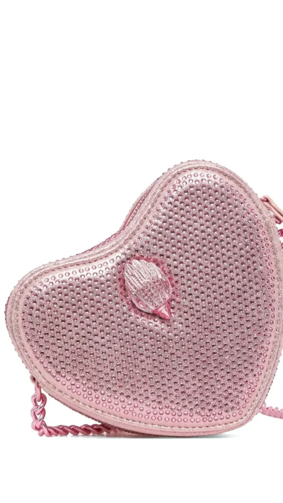 An image from file "4_18.webp" displaying a close-up, cropped product shot of a glamorous, heart-shaped crossbody handbag against a solid white background. The handbag is covered entirely in shimmering, metallic light pink crystals or rhinestones. Placed at the center of the heart structure is a signature, decorative metallic eagle head emblem in a matching pink tone, also encrusted with tiny crystals. A metallic pink chain link strap is visible attached to the lower-left corner of the bag, complementing its high-shine, feminine aesthetic.