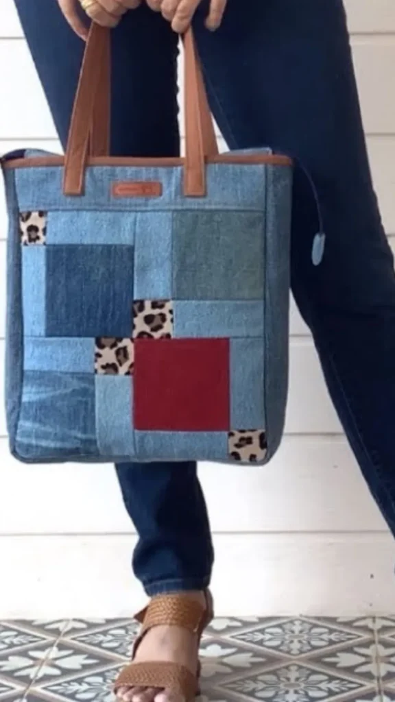 An image from file "4_19.webp" showing a person holding a handmade square denim patchwork tote bag. The bag features an intricate layout of various blue denim squares alongside a solid dark red fabric square and multiple smaller accents with a brown leopard print pattern. The upper border of the tote is finished with a thin brown trim, and it features two matching brown leather-like top handles. A small brown rectangular label is centered near the top edge. The person holding the bag is wearing dark blue jeans and brown woven slide sandals, standing on a grey and white geometric patterned tiled floor against a white paneled wall background.