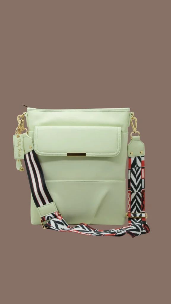 An image from file "4_20.webp" showcasing a light mint green faux leather crossbody laptop bag standing upright against a solid muted brown background. The bag features a large front pocket with a structured flap closure accented by a slim gold metal bar at the center. It includes a wide, detachable woven fabric guitar-style shoulder strap attached via gold lobster clasps. The strap is heavily patterned with a monochrome black, white, and red geometric chevron print on one section and bold stripes on another. A matching mint green tag reading "Pretty Pockets" hangs from a gold chain on the left side hardware.