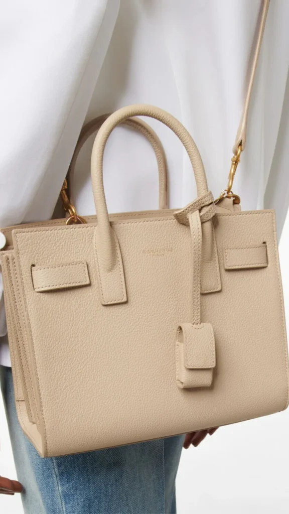 An image from file "4_21.webp" showcasing a close-up side view of a person carrying a beige Saint Laurent Sac de Jour handbag. The structured, boxy tote bag is crafted from finely grained beige leather and features rolled top leather handles, signature accordion pleated sides, and decorative leather straps looped through the front panel. A matching leather clochette containing a padlock hangs from one of the handle bases, and a faint gold embossed "Saint Laurent Paris" logo is subtly stamped near the top center. The bag is worn with an attached slim leather shoulder strap using gold-tone metal hardware. The person is dressed in a crisp white top and blue denim jeans.
