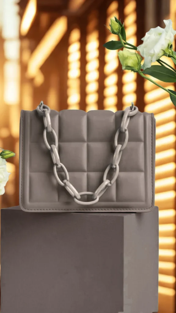 A structured product photograph from the file 4_10.webp showcases a rectangular, muted grey-brown handbag with a grid-like quilted pattern. The bag rests on a dark grey pedestal blocks and features a thick, matching plastic chunky chain strap draped across the front. White flowers with green leaves frame the top right and left corners of the composition. In the heavily blurred background, golden sunlight streams through window blinds, casting warm, horizontal lines of light.