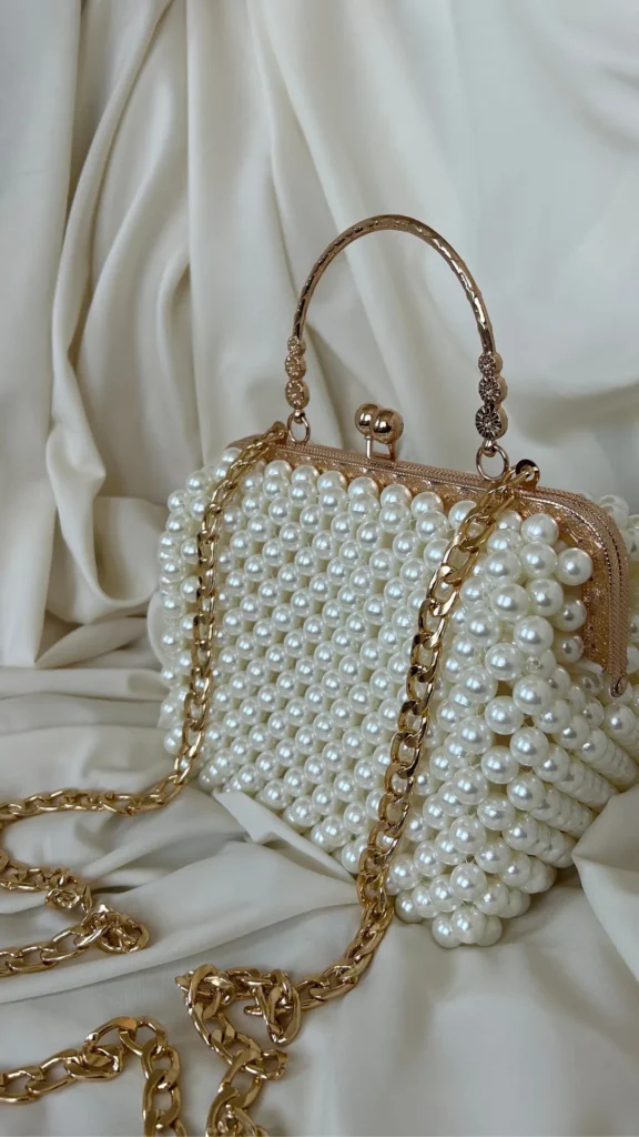 An image from file "4_13.jpg" showing an elegant, vintage-inspired evening clutch handbag completely covered in large, lustrous white faux pearls woven together in grids. The bag is designed with a structured rose-gold metal frame and a classic kiss-lock clasp at the top. It features a rigid, textured rose-gold metal top handle with decorative ornate detailing at the base, and a matching rose-gold curb chain shoulder strap draping forward. The entire bag rests on a background of softly folded, silky cream-colored fabric.
