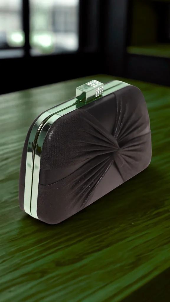A product photograph from the file 4.png shows an elegant, structured evening clutch bag resting on a polished green wooden surface. The clutch is crafted from a dark brown or charcoal satin fabric featuring a chic, central twisted ruching pattern that radiates outwards. It is framed by a sleek, polished silver metallic border and features a matching silver rectangular clasp set with small, glittering crystals on top. The background is softly out of focus, suggesting a dimly lit, sophisticated indoor venue.