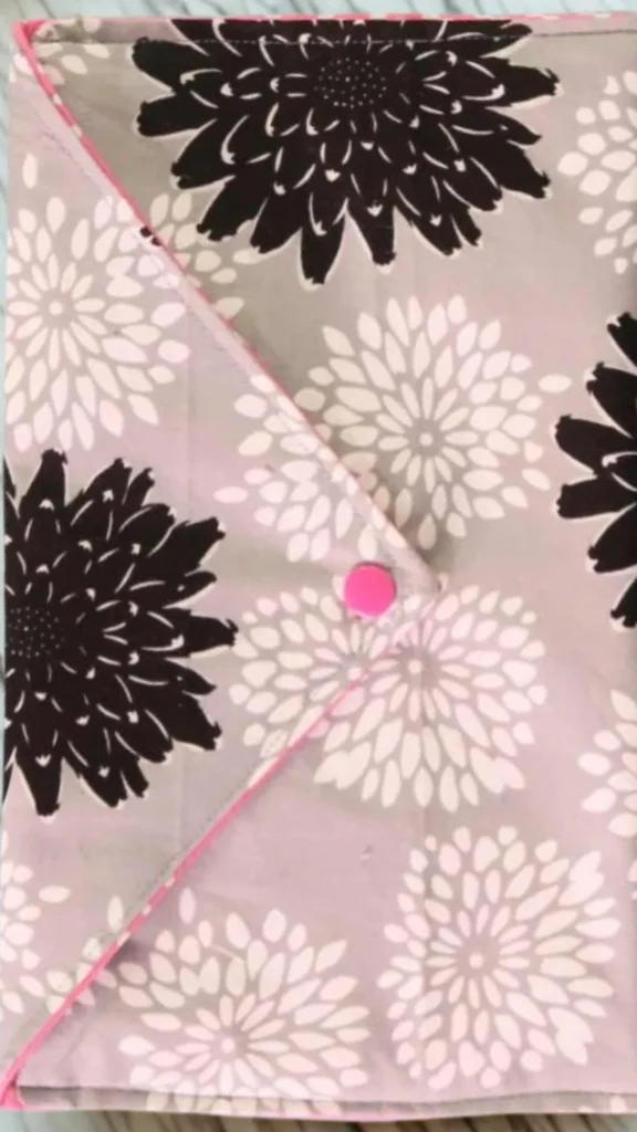 A top-down, close-up shot from the file 4_8.webp features a handmade envelope-style fabric clutch. The bag is crafted from a grey or muted lavender cotton fabric patterned with large graphic chrysanthemums or dahlia flower blooms in solid black and contrasting white. The bag has a distinct triangular fold-over flap with a thin pink stitched border that secures in the center with a bright pink snap button closure.