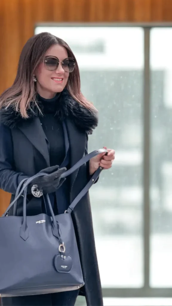 A woman smiling while holding a large navy blue leather tote bag outdoors in 5 (1)_2.webp. She is wearing a dark, elegant winter outfit consisting of a high-neck black top layered under a sleeveless black coat vest with a plush black faux-fur collar. She is accessorized with large tinted sunglasses, small dangling earrings, dark grey gloves, and the navy handbag which features thin top handles and a heart-shaped leather charm attachment.