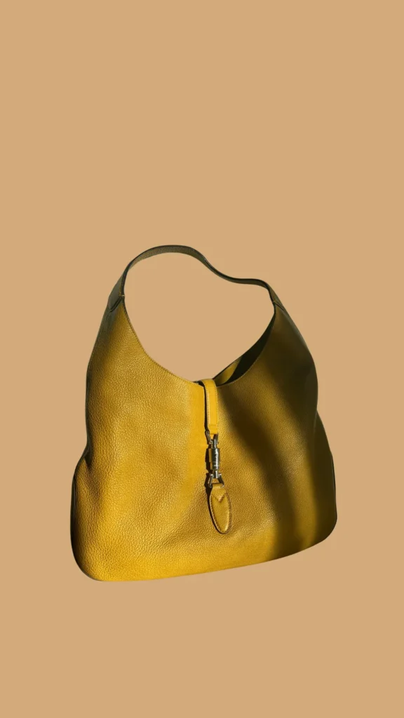 A product shot from 5 (1)_3.webp showcasing a vintage Gucci Jackie hobo bag set against a warm, muted tan background. The crescent-shaped handbag is crafted from a richly textured, pebbled leather in a vibrant mustard yellow hue. It features the signature curved hobo silhouette, a matching leather shoulder strap, and the iconic silver-tone metal piston clasp closure down the center front, cast in sharp shadow from a directional light source.