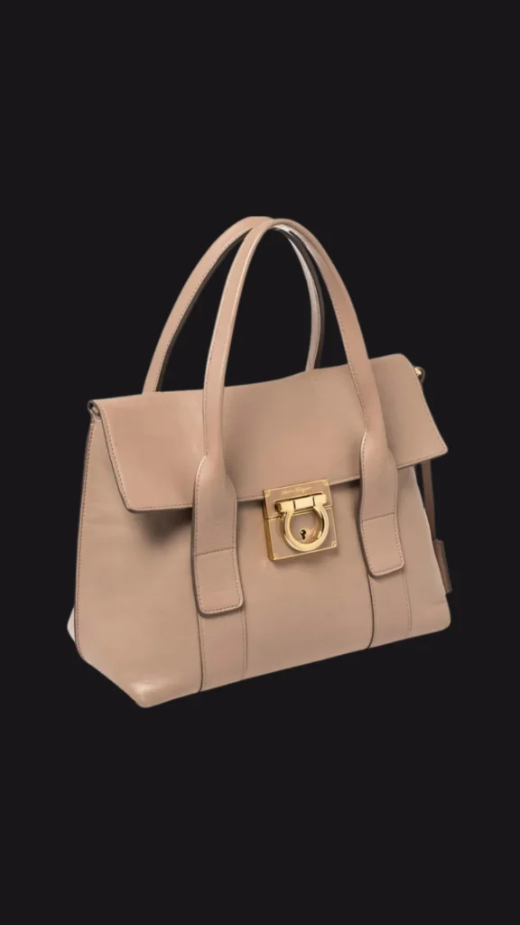 A product shot from 5 (1)_4.webp showcasing a structured, timeless beige leather satchel handbag against a solid black background. The bag features an elegant top flap design with twin structured top handles extending down the front panels. It is secured by a prominent, square gold-tone metal plate featuring a classic Gancini flip-lock clasp closure in the center, exuding a sophisticated "quiet luxury" style.