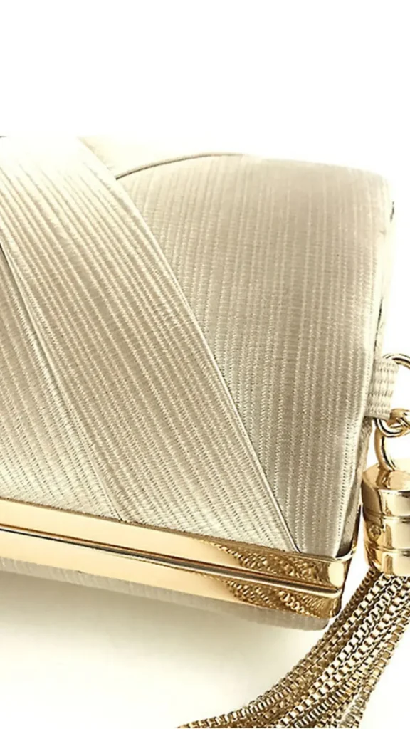 An image from file 5 (1)_3.webp showing a close-up, detailed view of an elegant, hard-case evening clutch bag. The bag is wrapped in a champagne-colored, finely ribbed satin fabric featuring an asymmetrical overlapping folded detail across the front. It has a polished gold metallic trim along the frame closure, and a substantial decorative gold chain tassel hanging from a hardware loop on the side.