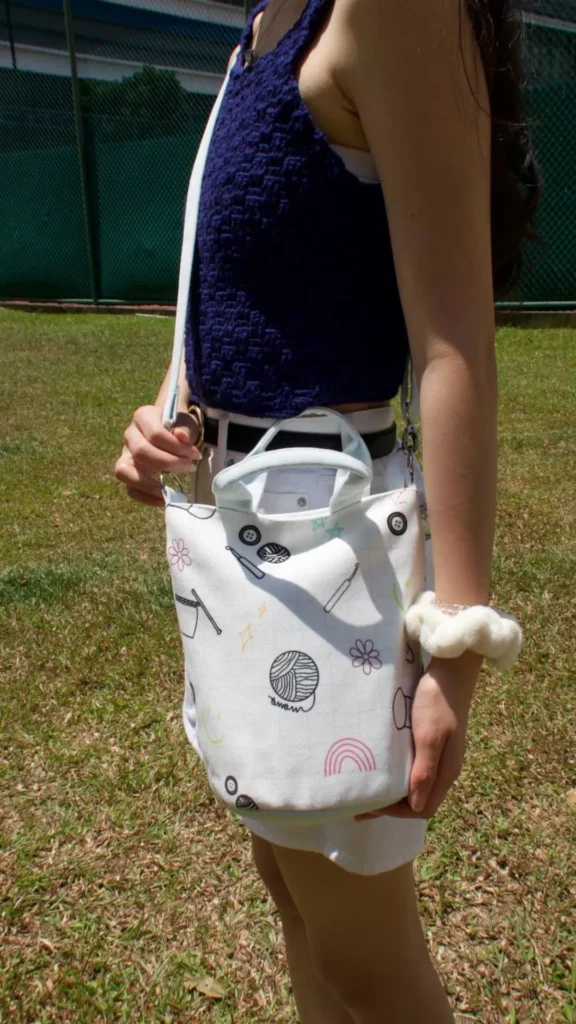 An image from file 5 (1)_4.webp showing a person standing outdoors on a grassy field, wearing a dark blue textured halter top and white shorts with a black belt. They are carrying a small, casual white canvas bucket bag adorned with playful, line-art sketches of knitting and sewing items like a ball of yarn, buttons, and a rainbow. The bag features short top handles and a long, light-colored shoulder strap slung over their shoulder.