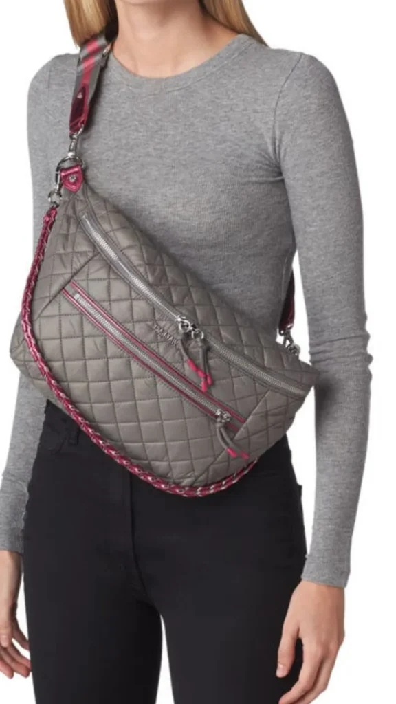 An image from file "5 (1)_6.webp" showing a close-up of a person wearing a large, casual quilted grey sling bag across their chest. The bag features a diamond-quilted pattern, multiple zippered front compartments with pink accents on the pull tabs, and a thick, adjustable woven shoulder strap. A decorative pink and white braided cord hangs along the bottom edge of the bag. The person is wearing a long-sleeved grey crewneck top and black pants against a plain white background.