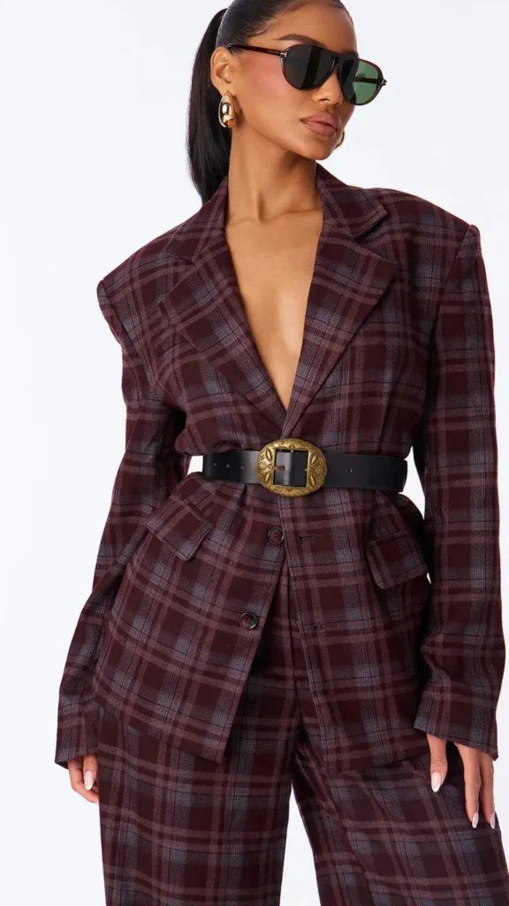A fashion model in 5_2.jpg posing in a matching maroon and dark grey plaid pantsuit against a white background. She is wearing an oversized blazer cinched at the waist with a thick black leather belt featuring a large, ornate gold buckle. The look is styled with dark aviator sunglasses and chunky gold hoop earrings, illustrating a playful yet powerful take on monochrome patterns.