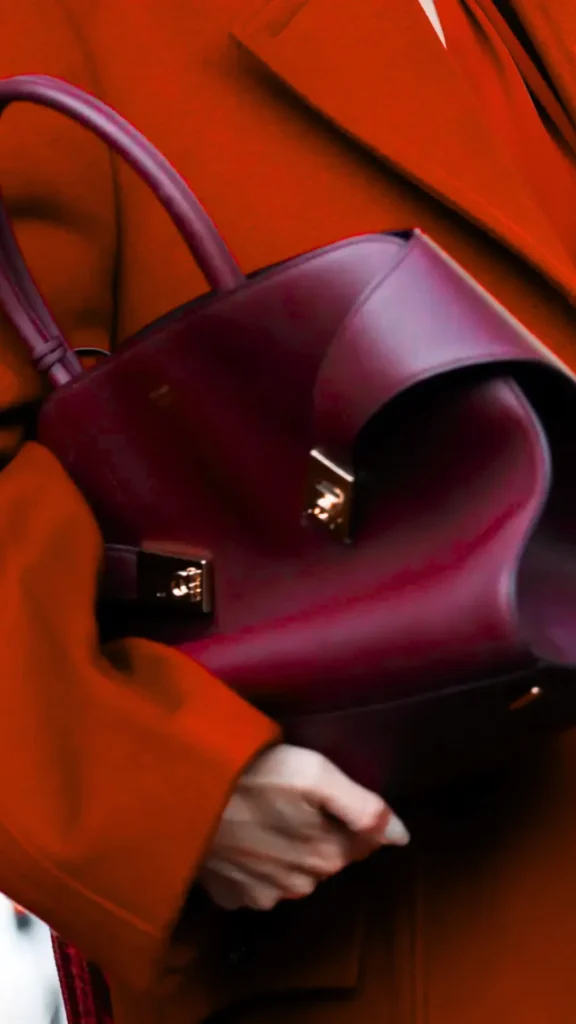 A dramatic, tight close-up shot focuses on a person's hand holding a luxury handbag. The bag is a structured, deep burgundy leather tote with a distinct curved top flap, smooth top handles, and sleek, minimalist gold metal clasps. It is being cradled against a vibrant burnt-orange or rust-colored wool trench coat, creating a bold, warm contrast. The framing highlights the textures of the fine leather and the fabric of the coat.