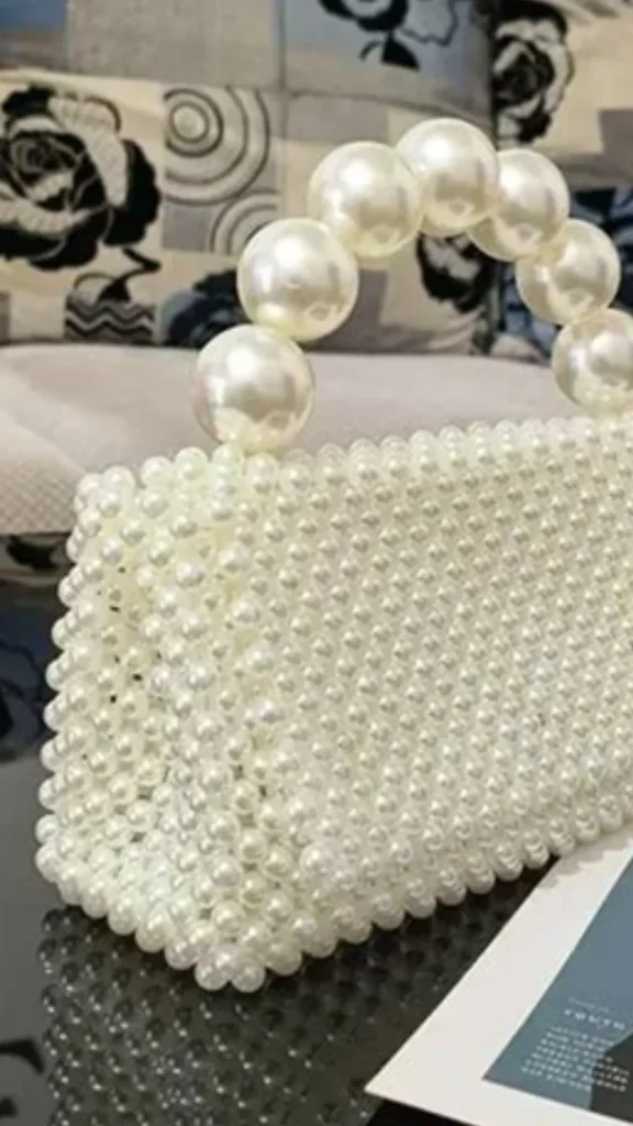 A close-up product photograph from 5_2.webp showcasing an exquisite, fully beaded white pearl handbag resting on a glossy black tabletop reflection. The body of the rectangular bag is intricately woven entirely from uniform, small glossy white pearl beads. It features a prominent, arched top handle constructed from a single row of massive, oversized round pearl statement beads. In the background, a soft fabric armchair with a grey and black floral pattern is visible.