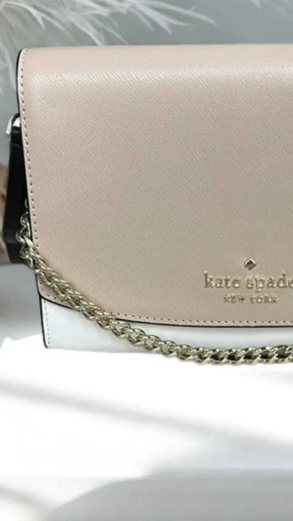 A close-up product photograph from 5_3.webp highlighting a structured Kate Spade New York handbag. The color-blocked crossbody bag features a cross-grain leather finish with a beige front flap closure, a white base, and a black side panel. The gold "kate spade NEW YORK" logo and signature spade hardware are embossed on the bottom right of the flap, complemented by a polished gold chain strap draped across the front.