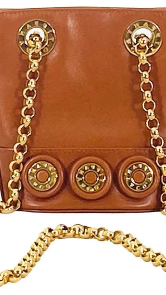 A close-up product photograph from 5_4.webp showing the detailed gold hardware on a brown leather handbag. The upper portion features two polished gold circular grommets with embossed lettering, through which a bright gold link chain strap is threaded. Along the lower stitched leather band, three decorative, circular gold button details are evenly spaced. An additional loose segment of the reflective gold chain strap rests in the foreground against a clean white background.