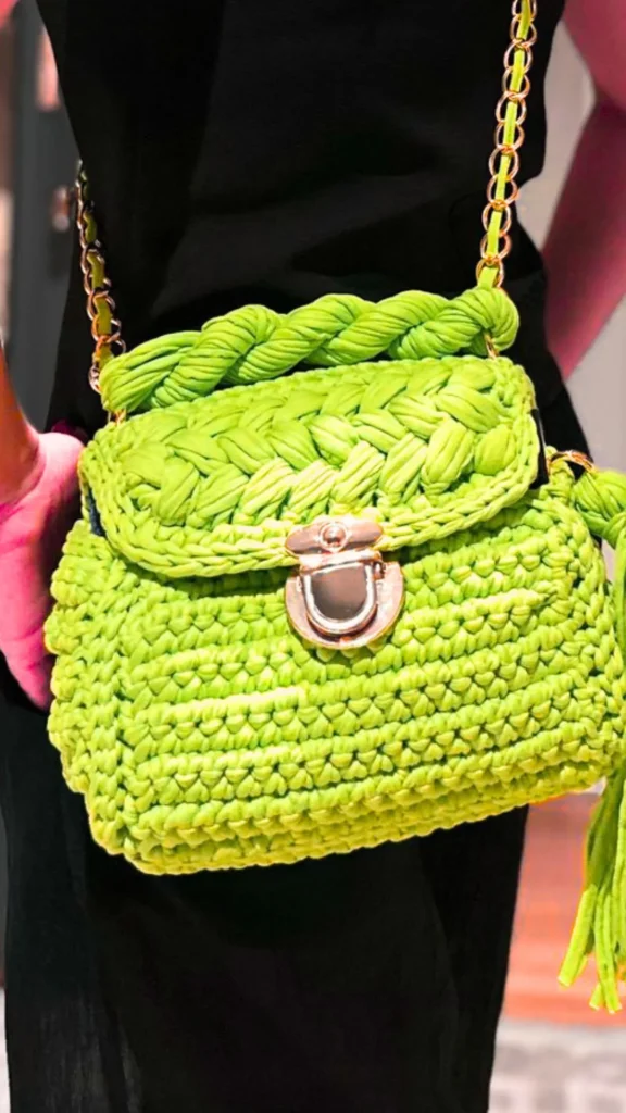 A close-up lifestyle photograph from 5_5.webp showcasing a vibrant pistachio lime green crochet handbag being worn by a person in a black outfit. The handmade bag features an intricate tightly-knitted yarn body, a thick braided top handle, and a large rounded silver metal push-lock clasp on the front flap. A matching green fabric tassel hangs from the side, complemented by a gold chain shoulder strap attached with gold metal rings.