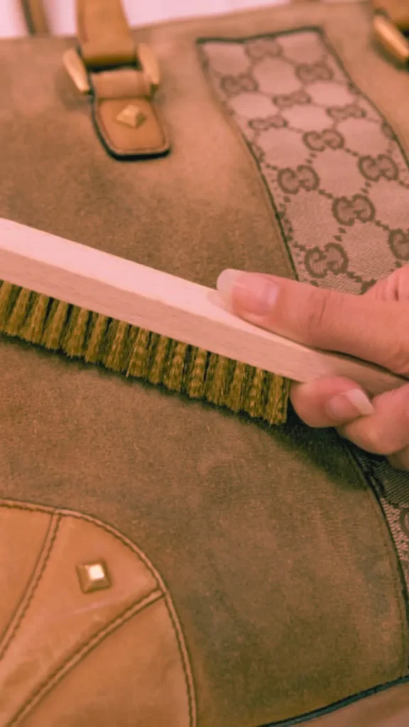 A close-up shot from the file 5_3.jpg shows a hand using a wooden-handled suede brush with coarse bristles to clean a green suede handbag. The luxury tote bag features tan leather panel accents with gold diamond studs at the bottom corner and a vertical canvas strip woven with a repeating designer monogram pattern down the side. The image has a warm, sepia-toned hue.