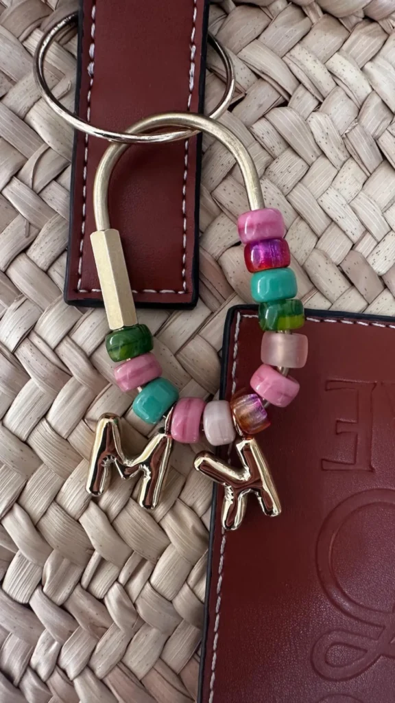 A close-up product shot from 5_7.webp showcasing a personalized initial bag charm attached to a woven straw tote bag with brown leather trim. The accessory features a large, gold-toned horseshoe-shaped metal ring threaded with an array of colorful, rounded beads in shades of pink, teal, green, and iridescent purple. Hanging from the center of the beaded loop are two puffy, polished gold letter charms forming the initials "M" and "K," adding a customized flair to the bag's hardware.