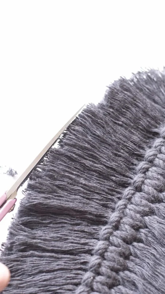 An action-shot photograph from 5_8.webp showing the process of crafting a DIY macramé feather or leaf bag charm. A pair of pink-handled scissors is shown carefully trimming the brushed-out, fringed edges of a dark grey cotton cord piece to create a clean, contoured shape. The center features a structured, braided knot spine, with the soft, dense, brushed yarn fibers fanning out flat against a white background.