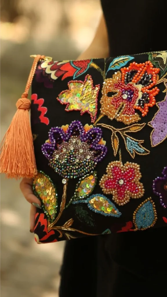 A close-up lifestyle photograph from 5_9.webp showing a person holding a black fabric zip-top clutch bag adorned with intricate, multi-colored floral embroidery and beadwork. The design features large stylized flowers and leaves completely filled with iridescent seed beads, sequins, and metallic threads in vibrant shades of orange, purple, pink, teal, and lime green. A long, peach-colored yarn tassel hangs from the gold top zipper, adding a final decorative touch against a soft, outdoor blurred background.