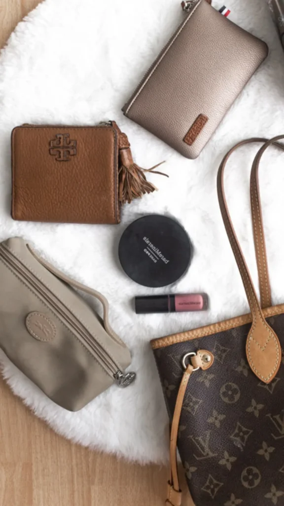 A flat-lay fashion photograph from 5_4.jpg showcasing a collection of premium personal accessories arranged neatly on a plush white faux-fur rug. In the bottom right corner sits a classic Louis Vuitton monogram coated canvas tote bag with tan leather straps. Surrounding it are everyday essentials for a mom's purse: a pebble-grained tan leather Tory Burch wallet with a decorative tassel, a champagne-metallic zip pouch, a beige nylon Longchamp cosmetic pouch, a round black bareMinerals makeup compact, and a small tube of pink lip gloss.