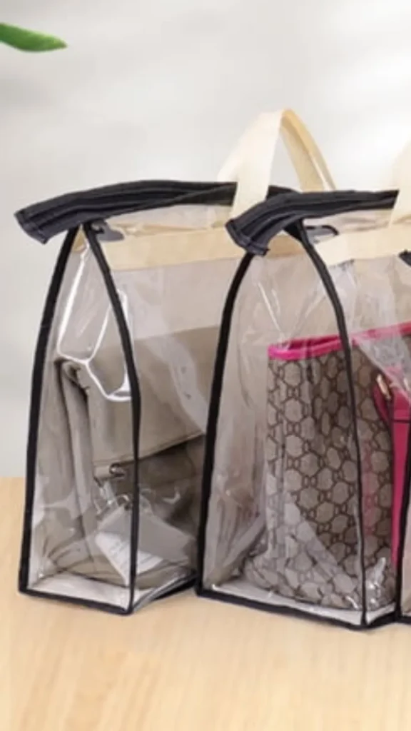 An image from file "5_16.webp" showing a set of two transparent plastic dust-proof storage organizer bags designed to keep handbags visible and protected. Each structured clear storage bag features a cream-colored top handle, black fabric trim along the seams, and a zipper closure. Inside the left organizer is a beige or grey leather folded crossbody bag, while the right organizer houses a structured brown patterned tote bag with bright pink interior lining and accents. The organizers sit side-by-side on a light-colored wooden surface.