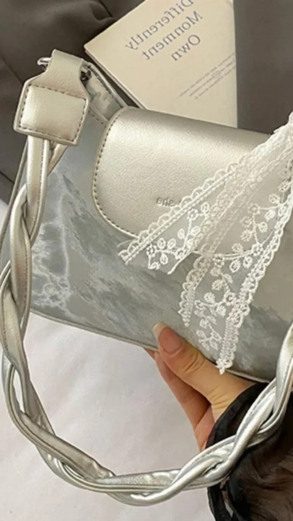 An image from file "5_17.webp" showcasing a close-up, flat lay styled photo of a metallic silver shoulder bag being held by a hand. The bag features a rectangular structure with a smooth silver front flap, a textured grey marbled pattern on the body, and a unique twisted or braided silver top handle. A delicate white lace ribbon with floral embroidery details is tied onto the front flap, adding a coquette, romantic touch to the modern accessory. In the blurry background, a book with its title printed in reverse sits next to dark fabric.