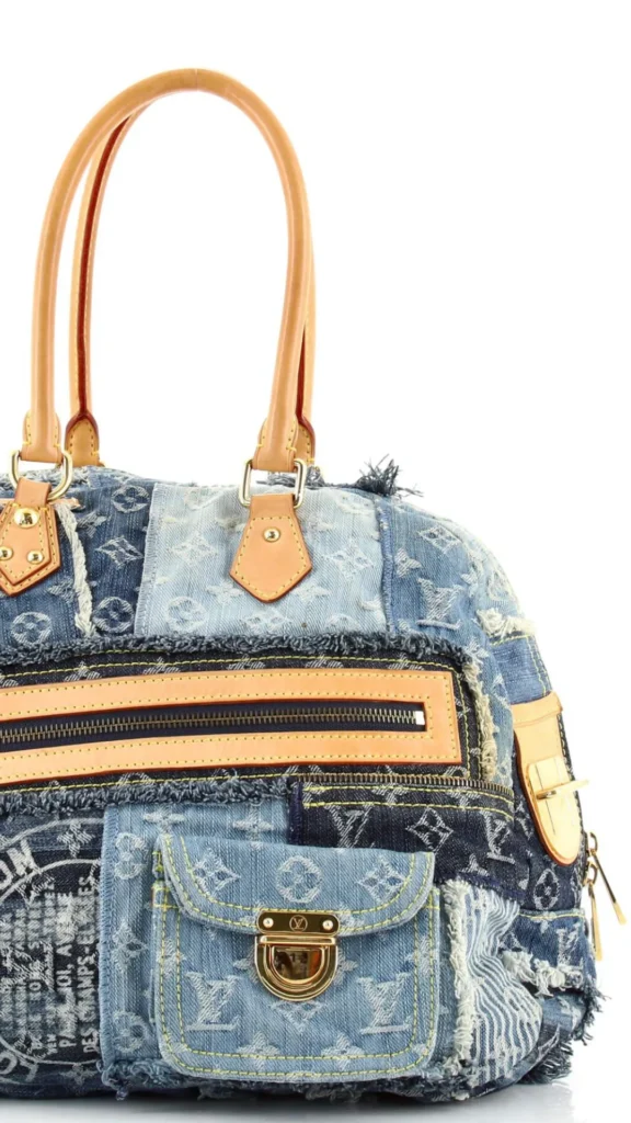 An image from file "5_18.webp" featuring a close-up shot of a luxury patchwork denim handbag against a plain white background. The bag is constructed from various shades of blue denim patches, heavily patterned with an all-over monogram logo print. It showcases intentional frayed and distressed raw edges along the seams. The handbag includes light tan leather top handles attached with gold hardware, a prominent horizontal zipper trimmed with tan leather on the front, and a lower flap pocket made of lighter blue denim with a gold clasp enclosure.