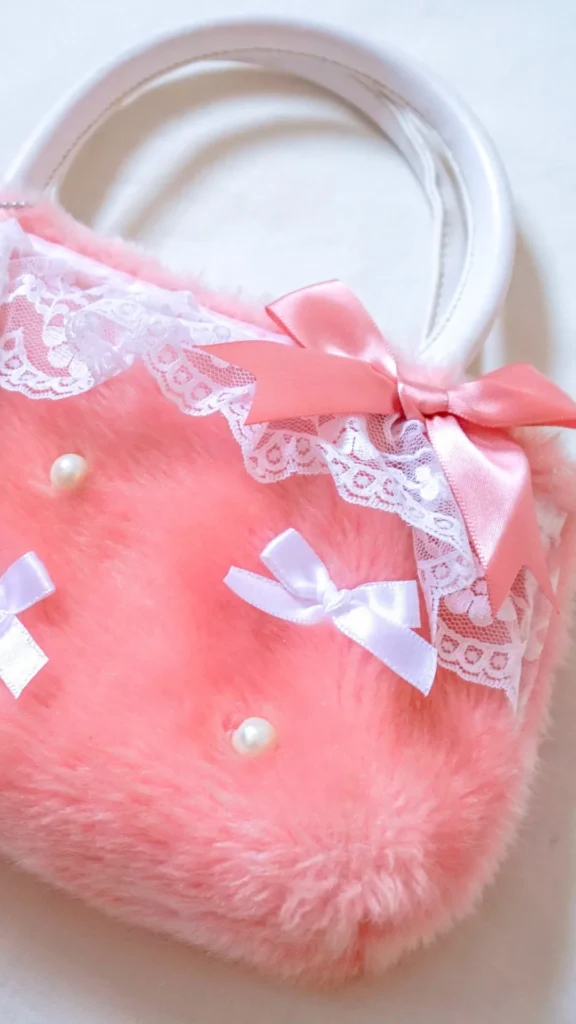 An image from file "5_14.jpg" showing a close-up of a whimsical, coquette-style handbag made from soft, fluffy pastel pink faux fur. The top trim of the bag is decorated with a layer of ruffled white lace and a matching pink satin ribbon bow near the handle base. The body of the plush purse is further embellished with small scattered white pearls and tiny white satin ribbon bows. The bag features smooth, curved white top handles and is captured against a plain white background.