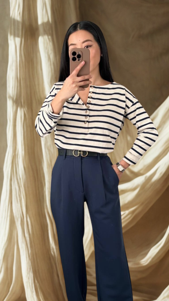 A mirror selfie from the file 5_9.png shows a person styling a classic, nautical-inspired outfit. The individual wears a long-sleeved cream top with horizontal navy blue stripes and a buttoned henley neckline, tucked into high-waisted, tailored navy blue trousers. The pants are accented with a black leather belt featuring a prominent gold double-gancini style clasp. The person holds a brown smartphone to block their face while posing with one hand in their pocket. The background consists of draped, textured cream or beige fabric backdrop.
