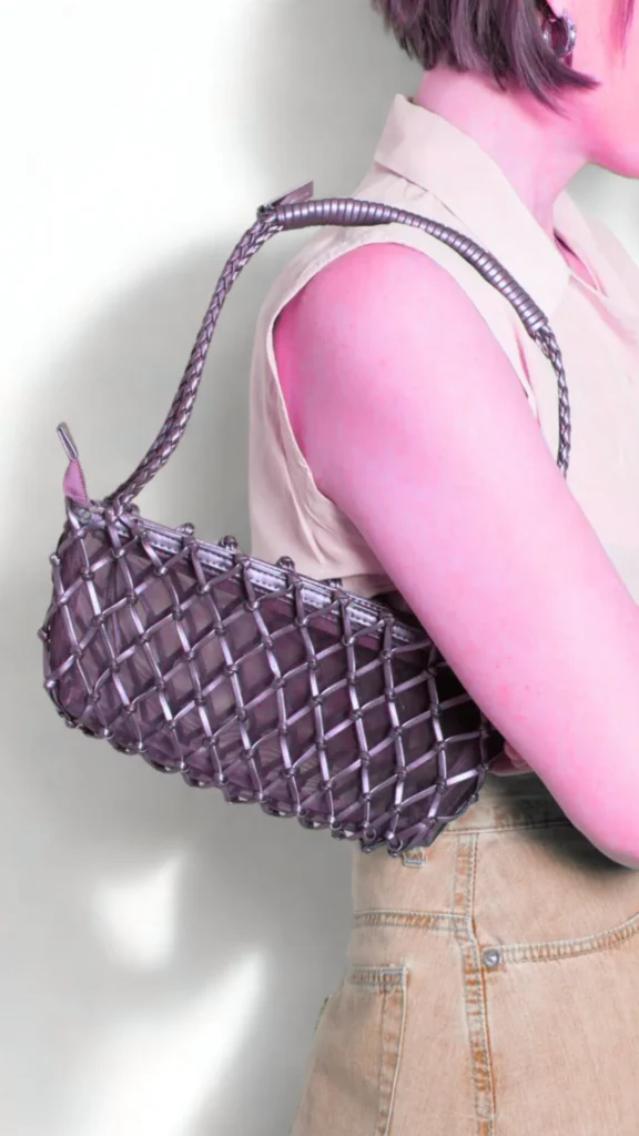 A close-up fashion photograph from the file 3.jpg highlights a structured, olive-bronze top-handle leather handbag. A hand holds the bag by its unique, rigid circular metal handle, which has a sleek gold finish. A matching leather tag with gold corner brackets and the embossed text "LMDS" hangs from a strap attached to the handle. The front of the bag features a small embossed logo depicting a kangaroo above the brand name.