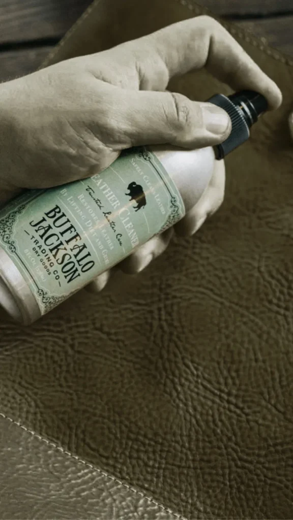 A close-up photograph from the file 5.webp shows a person's hand holding a spray bottle of "Buffalo Jackson Trading Co. Leather Cleaner" over a textured brown leather surface. The product label features a bison silhouette and vintage typography. The image focuses on the practical application of leather care and maintenance products.