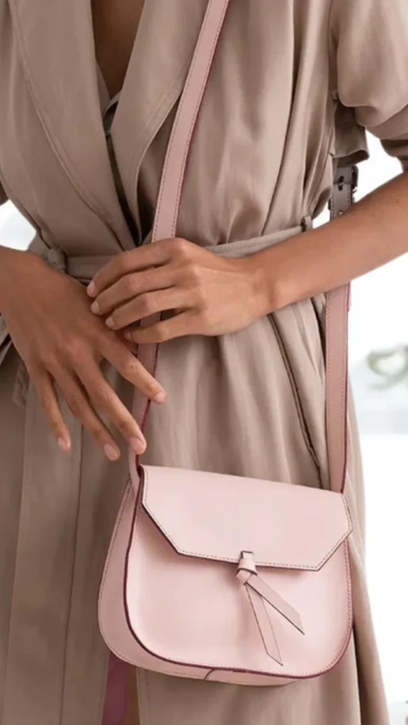 A close-up fashion photograph from the file 6.webp focuses on a person wearing a relaxed, belted taupe or beige trench coat. Slung over their shoulder is a structured, saddle-style crossbody handbag in a soft blush-pink leather. The bag features a clean, angular front flap accented by a minimalist, knotted leather tie detail hanging down the front.