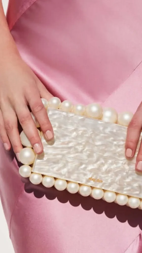 A close-up photograph from the file 6_5.webp captures a rectangular evening clutch bag being held in both hands against a smooth, light pink satin dress. The hard-case clutch features a glossy, iridescent white mother-of-pearl or acrylic marbled center surface. The entire border of the bag is framed by large, prominent ivory pearls of varying sizes. Tiny, subtle gold-toned text reading "may then" is stamped near the lower right corner of the clutch's face.