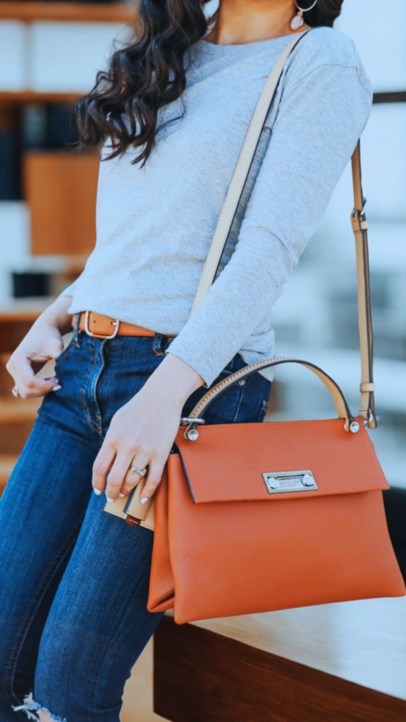 A close-up shot from the file 6_2.png shows a person leaning against a wooden surface, showcasing a smart-casual outfit and a prominent designer handbag. The individual wears a plain, long-sleeved light grey top tucked into dark blue jeans accented with a tan leather belt. Slung over their shoulder is a structured, vibrant orange-cognac leather top-handle satchel bag, which features a rectangular front flap and a horizontal silver metal clasp closure. A thin, beige leather shoulder strap extends upward, and the blurred background suggests a modern indoor setting.