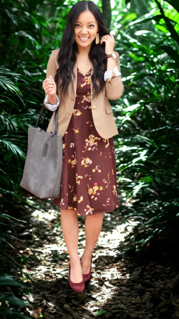 A smiling woman with long wavy dark hair standing on a path surrounded by lush green foliage in 6_2.png. She is wearing a maroon V-neck midi dress with a yellow floral pattern, layered under a tan tailored blazer with gold buttons. The outfit is completed with a large grey leather tote bag, dark maroon suede pumps, and tan dangling earrings, illustrating a versatile day-to-night smart casual aesthetic.