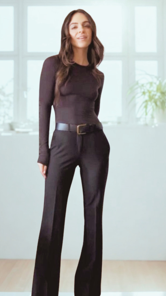A full-length shot from the file 7_2.png features a woman with long, wavy brown hair posing in a sleek, monochromatic outfit. She wears a form-fitting, long-sleeved black top tucked neatly into matching high-waisted, slightly flared black trousers. Her waist is defined by a wide black leather belt with a square, antique-gold buckle. She stands in a brightly lit room with a light wood floor, set against a blurred background showing a large window and a green houseplant.