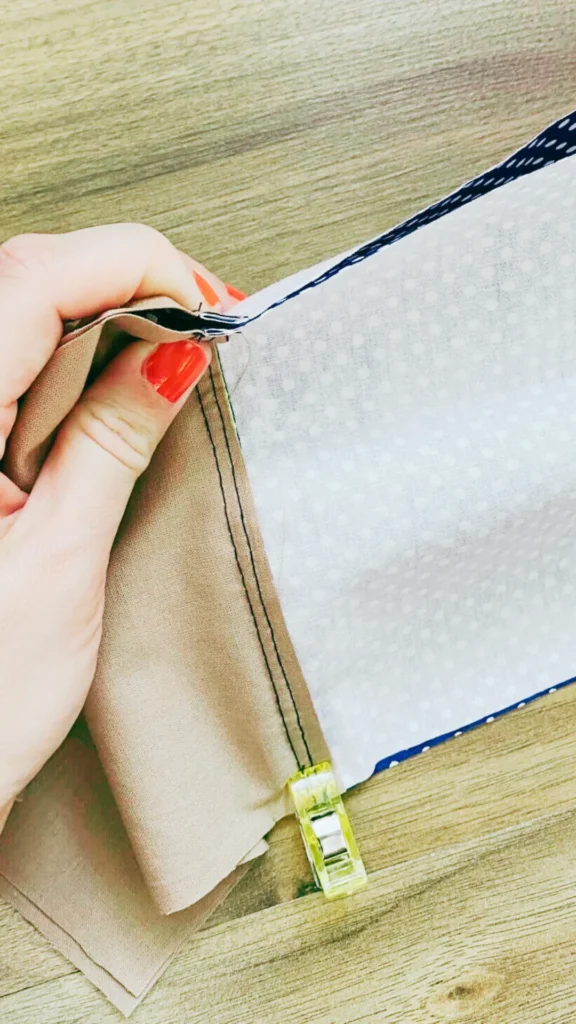 A top-down, close-up shot from the file 8.webp shows a person's hands with bright orange-red nail polish holding together pieces of fabric during a sewing project. The work-in-progress reveals a tan or beige fabric layered next to a white fabric lining with a faint dot pattern, showing a dark stitched seam along the edge. A small, translucent yellow plastic sewing clip secures the fabrics together at the bottom of the seam. The entire project rests on a light-colored wooden table or work surface.