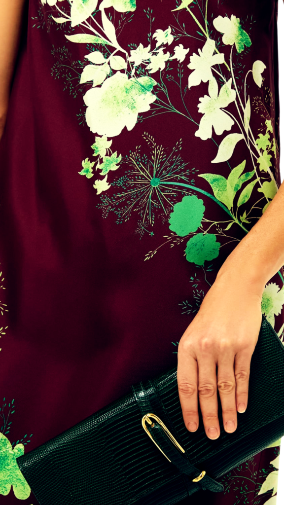 A close-up shot from the file 9.jpg shows a person holding a sleek evening clutch bag against a floral dress. The clutch is crafted from deep emerald green faux-lizard or faux-croc textured leather, featuring a horizontal rectangular shape and a vertical leather tab closure secured with an elongated, oval-shaped gold metal buckle. The person's hand rests over the top of the bag. They are wearing a sleeveless silk or satin shift dress in a dark maroon or burgundy hue, decorated with a vibrant graphic print of pale mint green and emerald green wild floral bouquets and delicate botanical illustrations.
