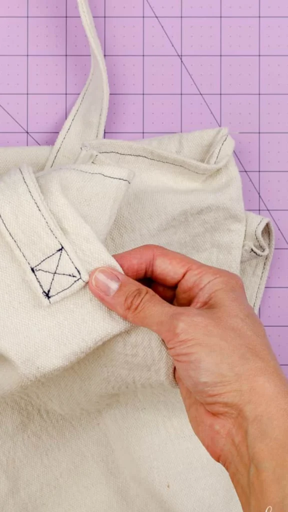 A close-up view of a hand holding the strap of a handmade off-white canvas tote bag. The strap is attached to the top edge of the bag with a visible box-and-cross reinforcement stitch using dark contrasting thread. The canvas fabric is folded over slightly, and the entire project rests on a purple cutting mat with grid lines.