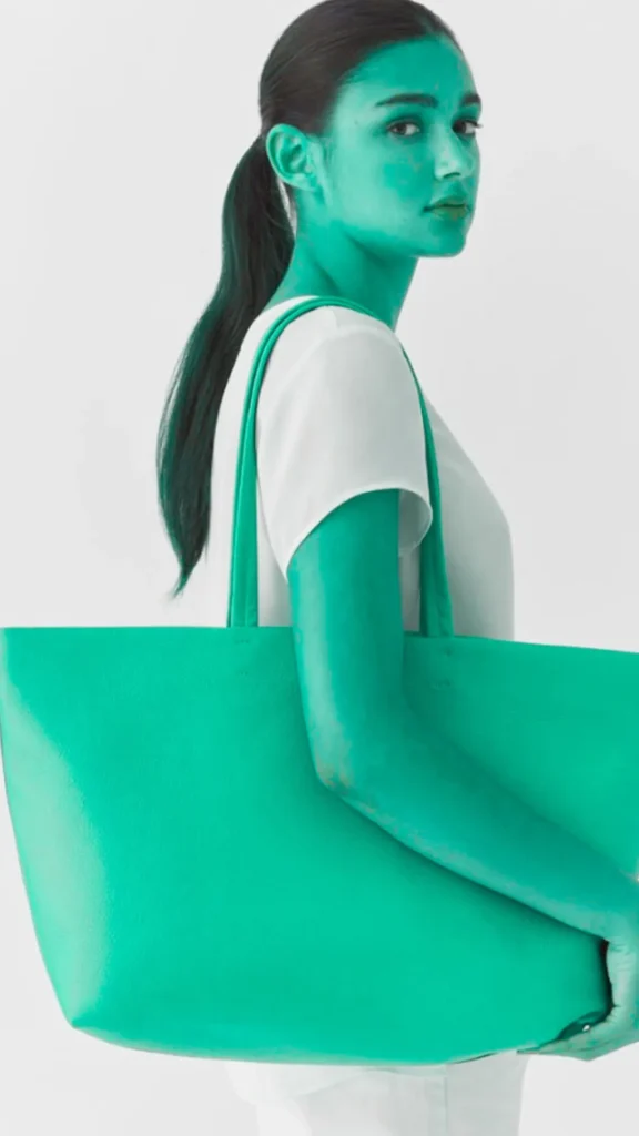 A woman stands in profile against a plain white background, wearing a simple white short-sleeved shirt. She has dark hair tied back in a ponytail and carries a very large, oversized teal-green leather tote bag over her shoulder, holding the base of the bag with her hand.