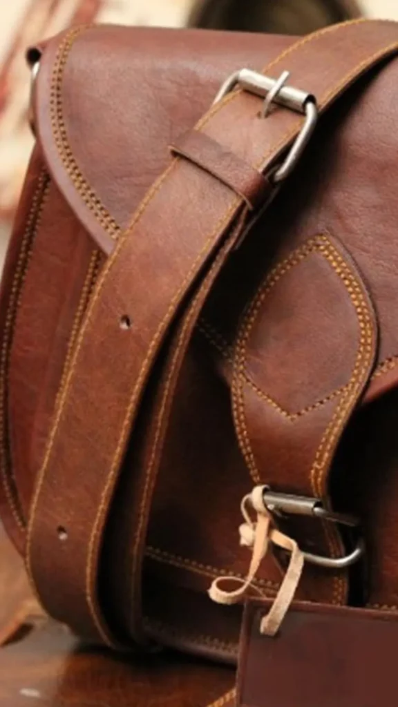 A close-up shot of a dark brown distressed leather satchel bag. A thick matching leather crossbody strap with pre-punched holes and a silver metal buckle drapes diagonally across the front flap. The satchel features prominent tan-yellow stitching outlining a pointed accent tab on the front pocket closure. A small piece of light-colored string is tied to the lower metal buckle clasp, highlighting the bag's textured, vintage school-bag aesthetic.