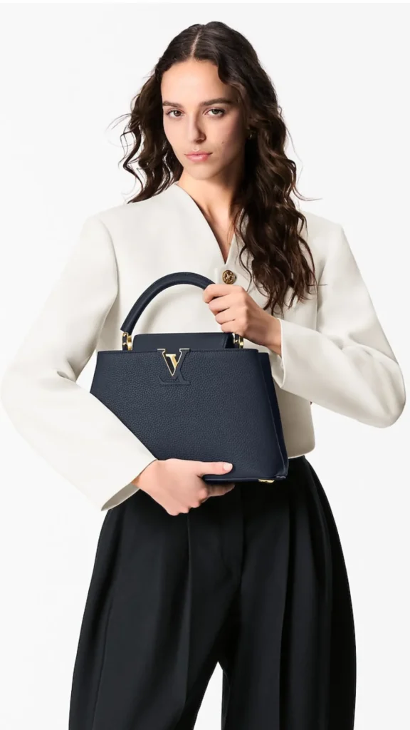Studio portrait of a woman with dark wavy hair posing against a white background. She is wearing a structured cream-colored blazer jacket with a prominent gold button and pleated black high-waisted trousers. She holds a structured navy blue Louis Vuitton Capucines handbag featuring textured grained leather, a curved top handle, and a gold metal LV initials logo accent prominently displayed on the front.