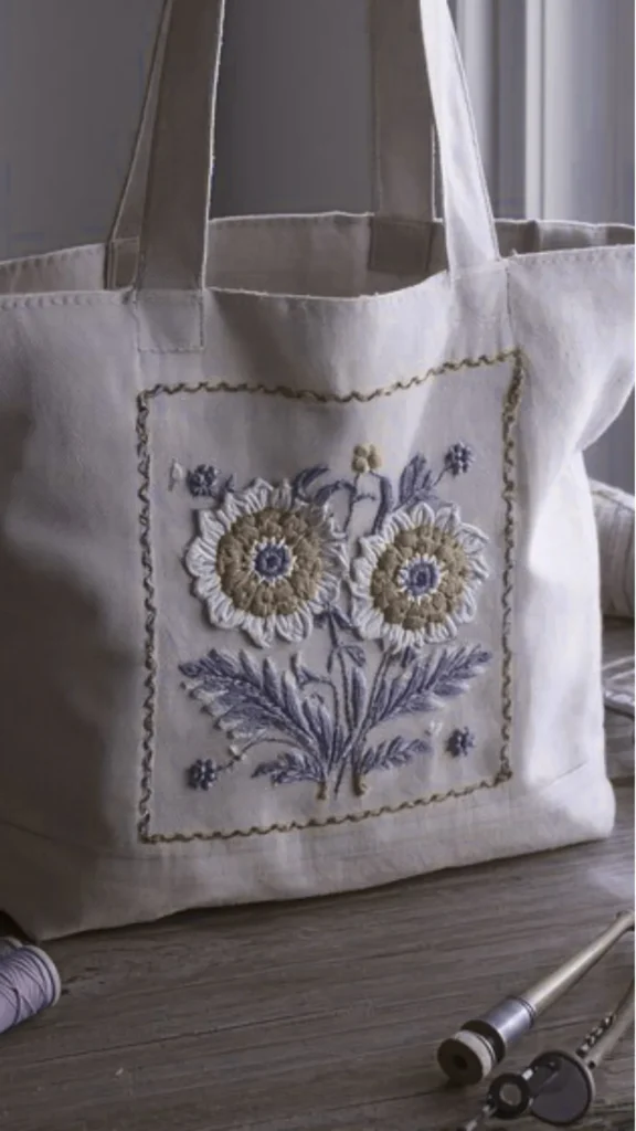 A close-up shot of a cream-colored canvas tote bag resting on a rustic wooden surface. The front of the tote features a large, square embroidered patch displaying a detailed floral pattern with two prominent white and beige sunflowers, muted purple leaves, and small accent blossoms, all enclosed by a decorative stitched border.