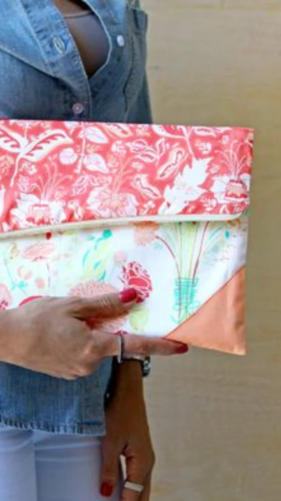 A person wearing a denim button-down shirt holds a handmade envelope-style fold-over clutch bag. The clutch features a bright coral and white floral pattern on the top flap, a lighter white fabric with pastel floral illustrations on the main body, and a solid peach-colored corner accent at the bottom. The person's hand with red nail polish is visible holding the bag against a neutral wall backdrop