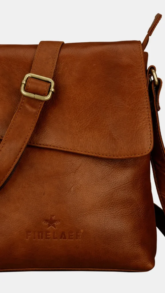 Close-up shot of a rich brown leather crossbody bag with a large front flap against a light background. A thick leather shoulder strap with a dull gold metal adjustment buckle runs diagonally across the front of the bag. The lower portion of the smooth leather surface features a debossed brand logo reading "FIDELAER" underneath a small star and bird emblem. Fine, matching brown stitching details outline the edges of the bag's flap and strap.