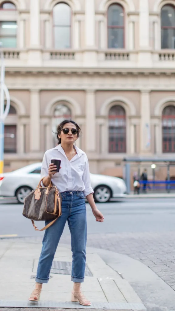 A woman with dark hair and sunglasses stands on a city street corner holding a coffee cup. She wears a crisp white button-down shirt with rolled sleeves, high-waisted medium-wash blue jeans with cuffed hems, and nude strappy block heels. On her arm, she carries a Louis Vuitton Speedy Bandoulière bag in the classic brown monogram canvas. An old European-style building with arched windows serves as the soft-focus background.