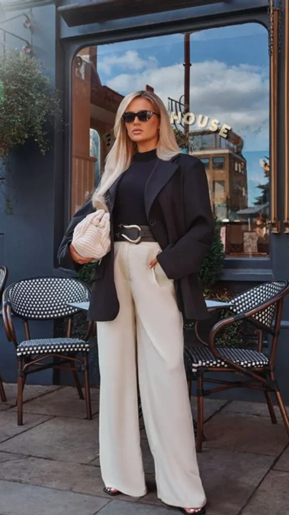A blonde woman standing outdoors in front of a cafe wearing high-waisted, wide-leg cream trousers, a black turtleneck top, and an oversized black blazer. She accentuates her waist with a wide black leather belt featuring a large gold buckle and wears black sunglasses. Under her arm, she carries a soft, ruched cream pouch clutch bag. Wicker cafe chairs and a glass storefront form the background behind her.